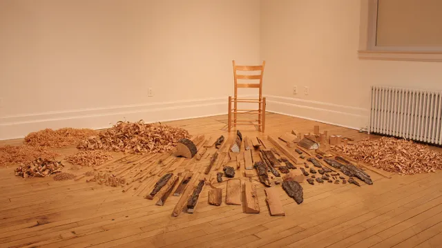 Interior of a museum, white walls and wood floors. In the corner a simple wooden chair and in front of it scraps of wood and wood shavings neatly piled. Art installation by Gina Siepel. 