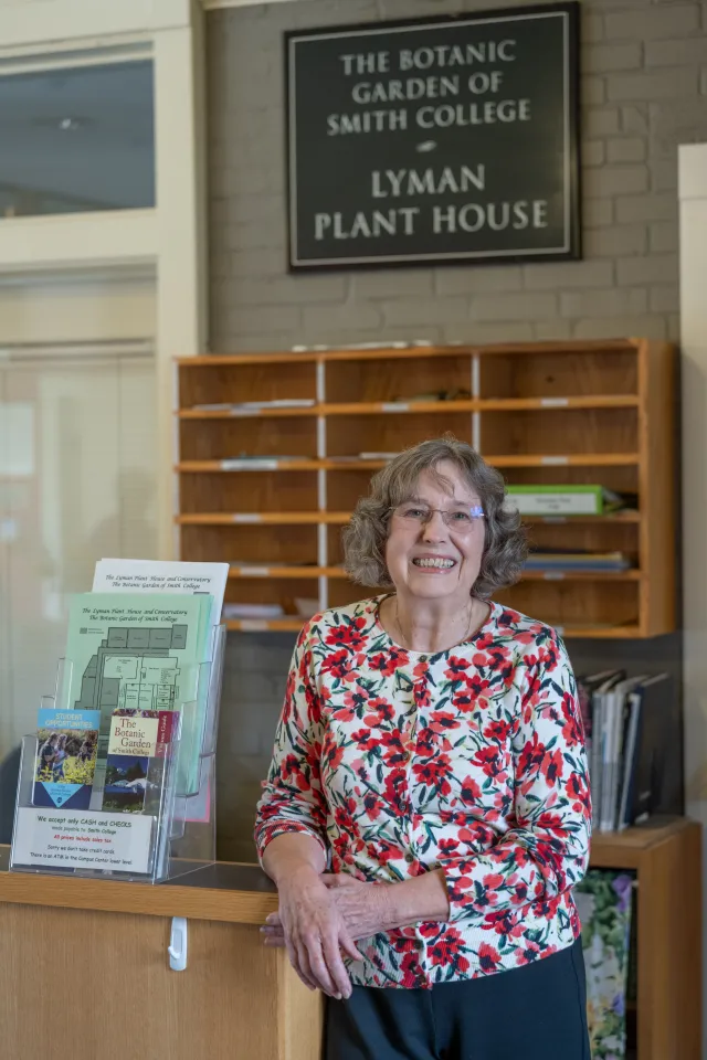 older woman with gray hair, glasses and a flowered red cardigan standing at a counter at the botanic garden lobby
