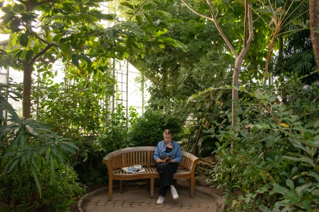 student in blue denim long sleeve shirt, black pants and white shoes on bench in a greenhouse surrounded by plants