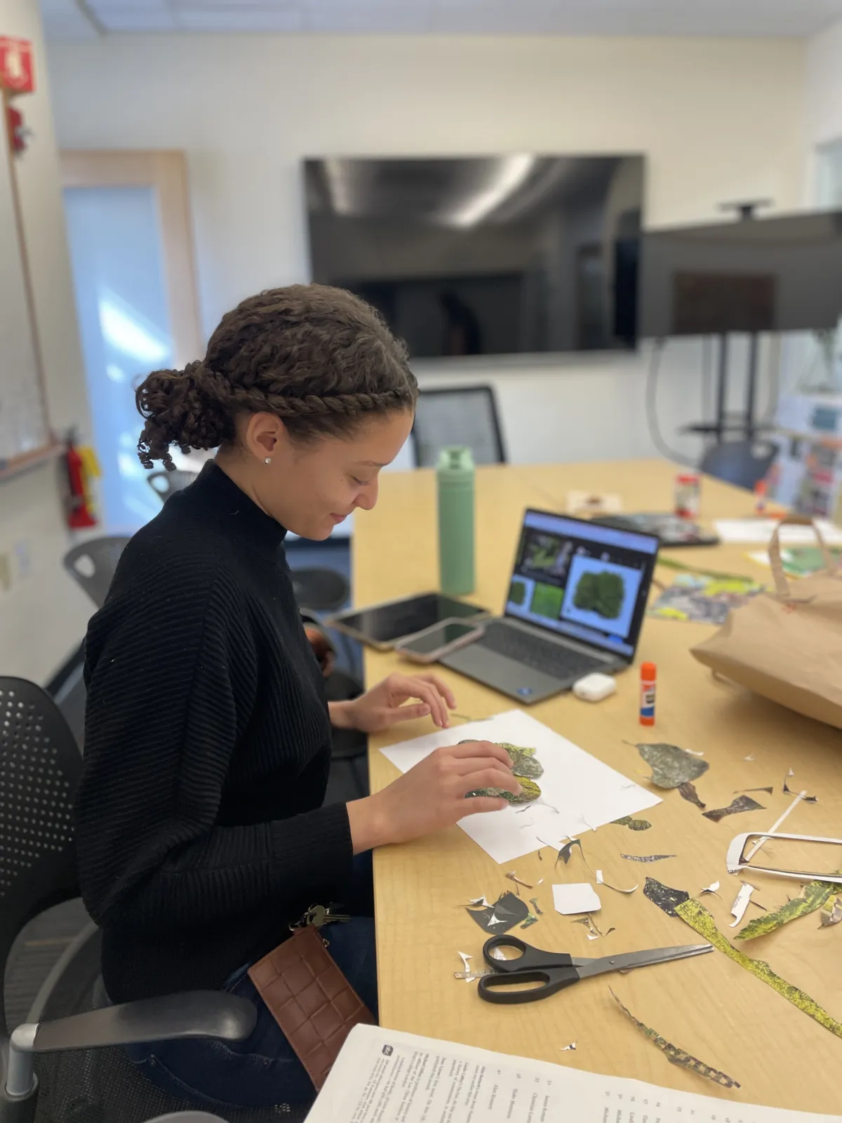 Student with brown hair and black turtleneck at a conference room desk working on a collage. 