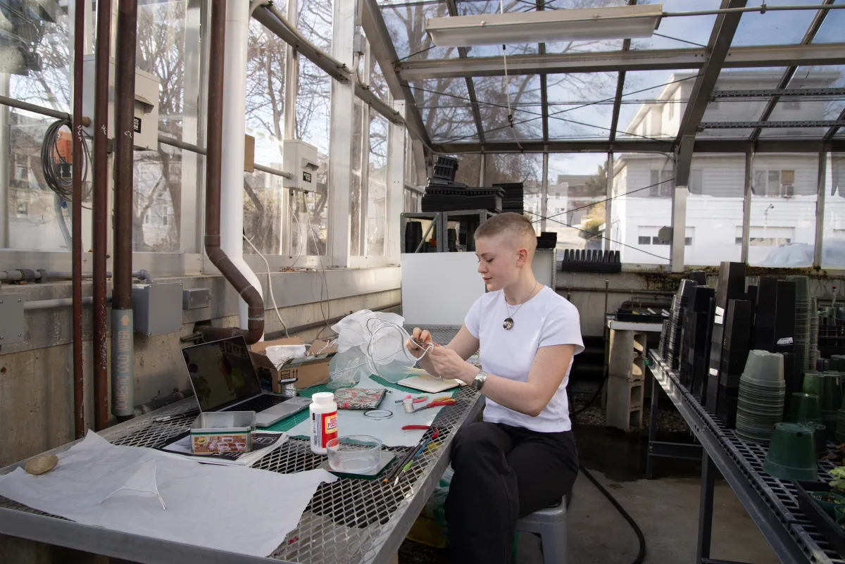 woman in white shirt and black pants in greenhouse working on a sculpture.